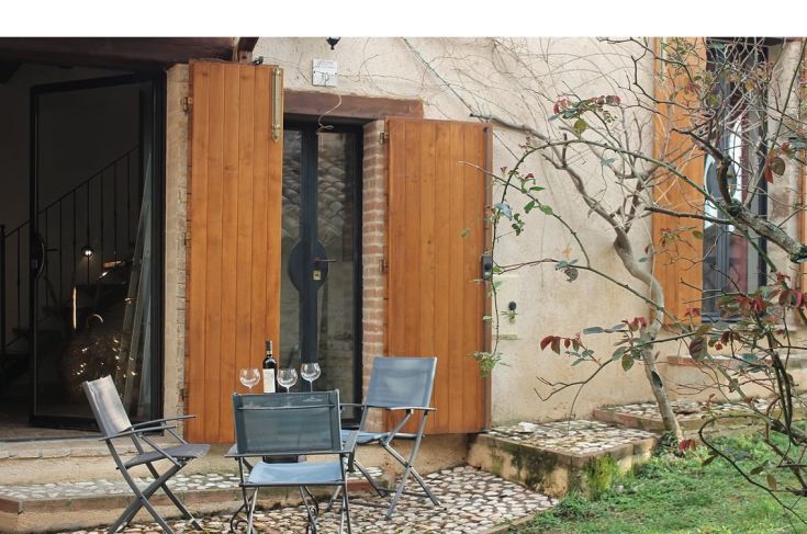 A portrait of an outdoor area featuring a table and chairs, with a bottle of wine, set in a historic context.