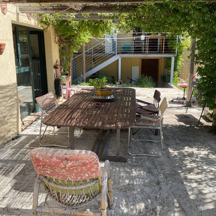 Ein einfacher Garten mit einem Holztisch und Stühlen, umgeben von Gras und Pflanzen. Ein ruhiger Ort.