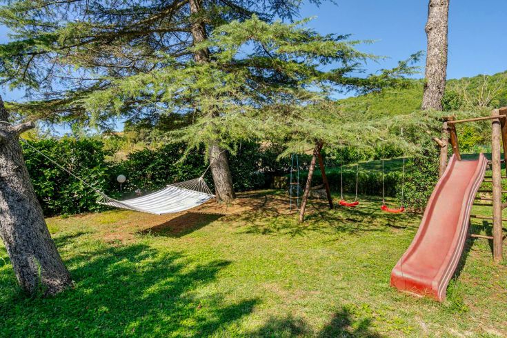 An outdoor space with children's play equipment, including swings, a slide, and a hammock, surrounded by greenery.