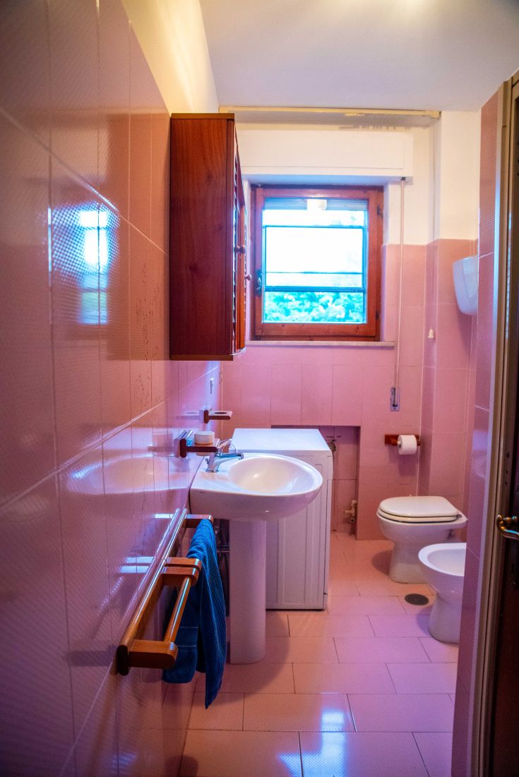 Bagno semplice con piastrelle rosa e dettagli in legno, dotato di una finestra che porta luce naturale.