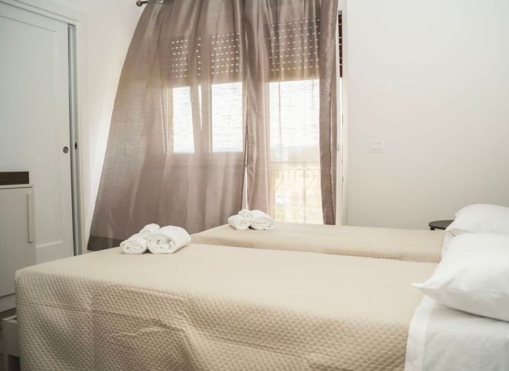 Chambre lumineuse avec des lits simples, des rideaux clairs et un mobilier simple.