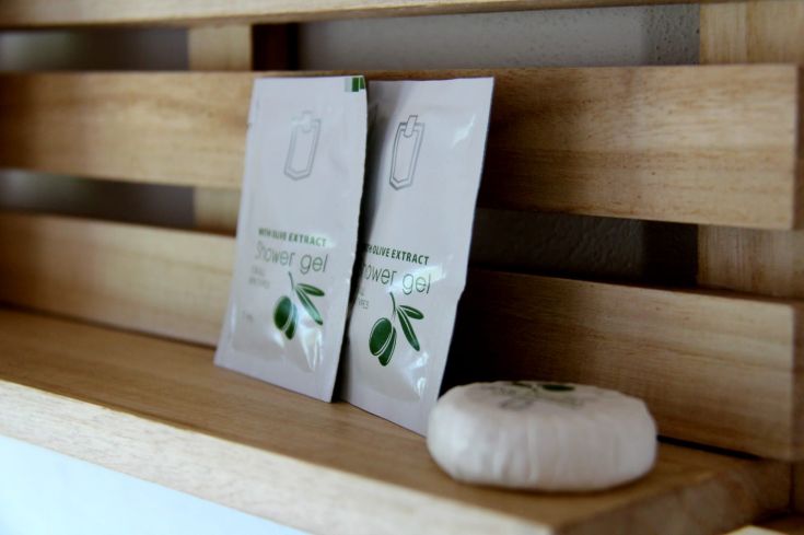 Due bustine di gel doccia con estratto di oliva e un sapone, poggiate su uno scaffale in legno.