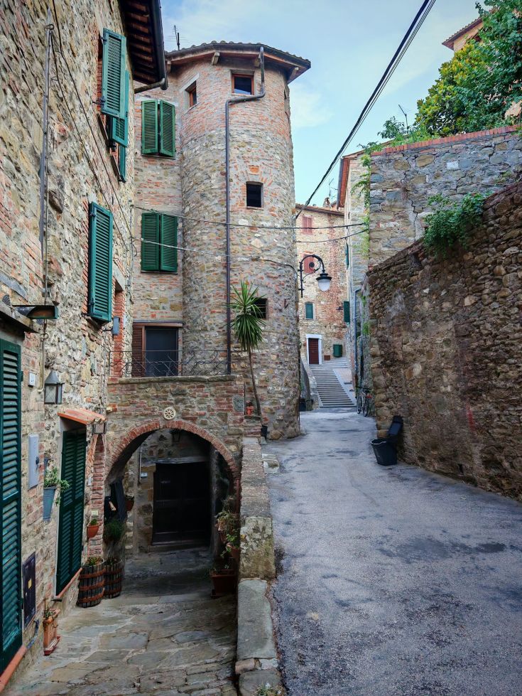 A narrow street in a historic village, featuring stone buildings and a tower in the background.