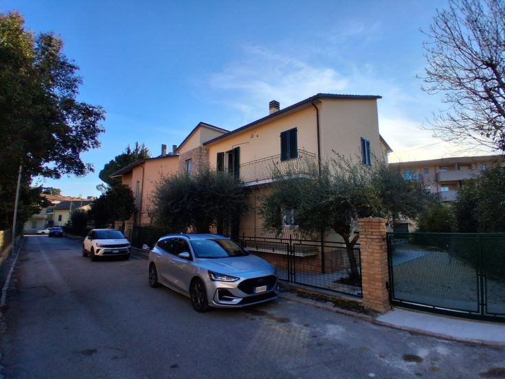 Villa surrounded by a large garden, with cars parked along a simple and quiet road.