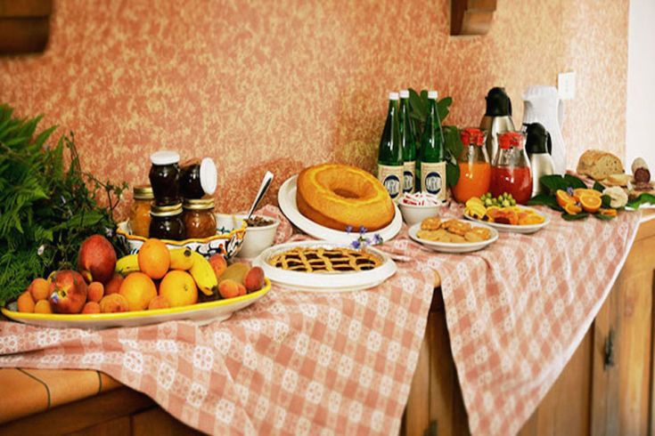 Su un tavolo sono disposti frutta fresca, dolci e bevande in un ambiente semplice e accogliente.