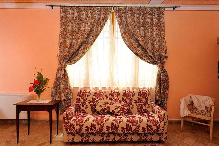 Un soggiorno semplice con divano a fiori e tende leggere, illuminato da finestre che si aprono su un giardino verde.