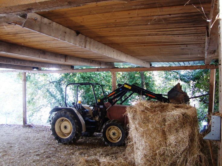 Un tracteur utilise un bras mécanique pour soulever des balles de foin à l'intérieur d'un bâtiment en bois.