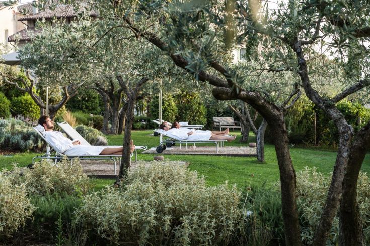 Two men in bathrobes relax in a well-kept garden surrounded by olive trees.