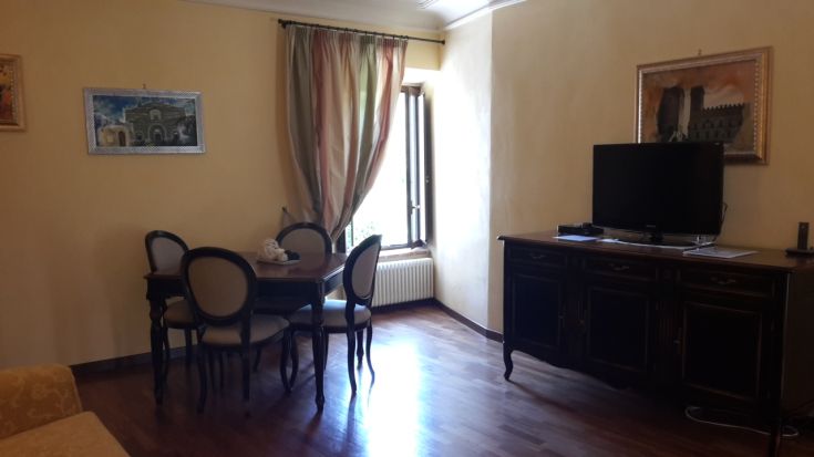 Ein schlichtes und gemütliches Wohnzimmer mit Tisch, Fernseher und großen Fenstern, die den Raum erhellen.