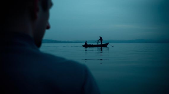 Silhouette of a traveler looking at the foggy lake horizon from a traditional boat at dawn.