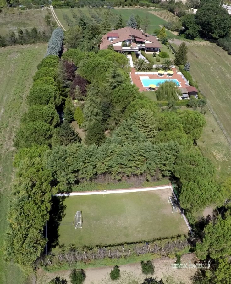 Ripresa aerea di un palazzo con piscina, circondato da alberi e un campo da calcio.
