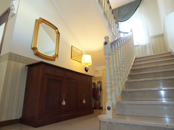 Un ingresso sobrio di una villa con scale, un lampadario e un grande specchio decorativo.