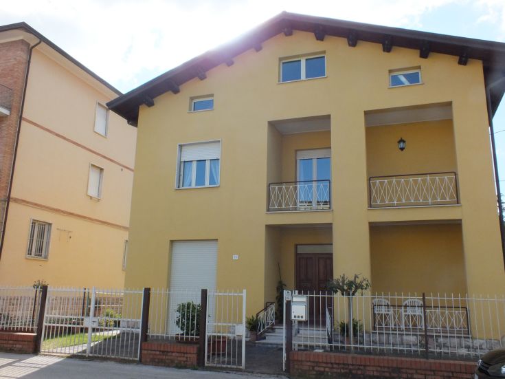 Una casa moderna gialla con un portico e balconi. Si trova in un'area verde ben curata, con un giardino ordinato.