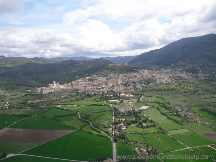 Panorama di Assisi con campi verdi e nuvole che sovrastano la città.