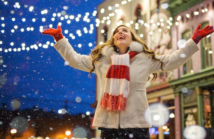 Una giovane donna sorride mentre gioca nella neve, circondata da luci decorate per le festività, godendo dell'atmosfera invernale.