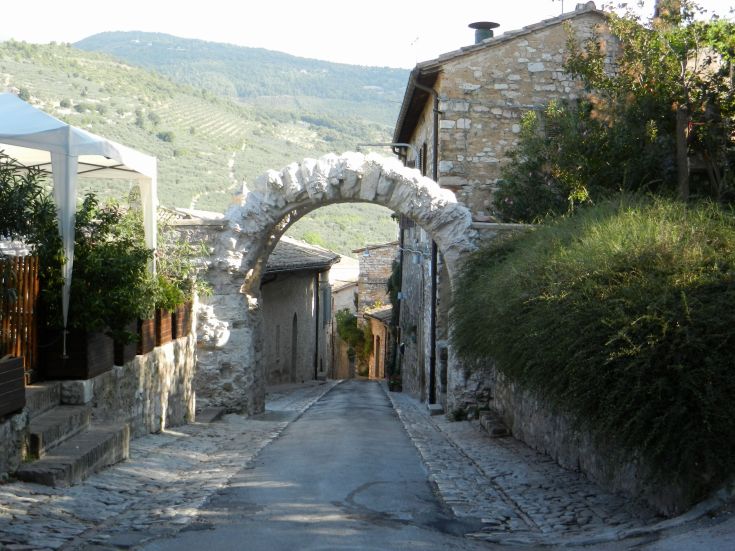 Strada in pietra con un caratteristico arco in pietra e palazzi storici in Umbria, circondata da alberi e colline.