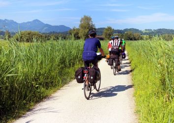 Ciclisti su un sentiero tra prati e boschi, circondati da colline e montagne verdi.