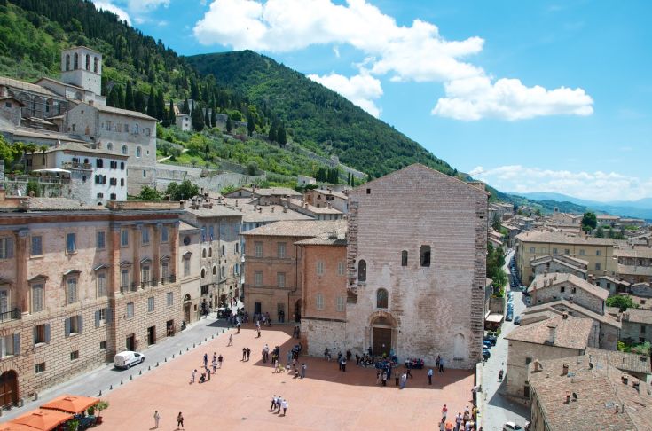 Una scena di una piazza con edifici storici, circondata da colline verdi.