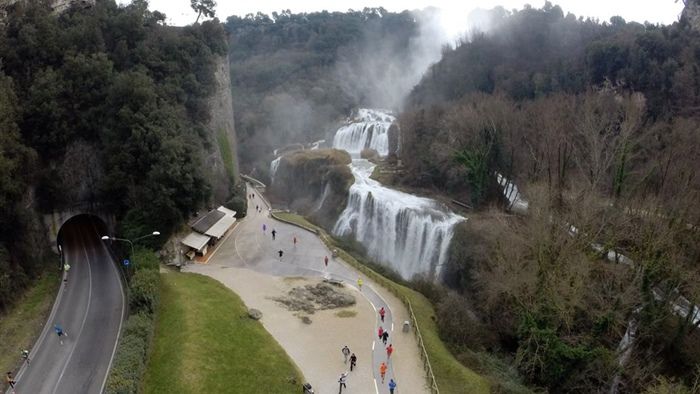 Una scena di una cascata circondata da vegetazione, con sentieri che si snodano tra gli alberi e visitatori. La cascata scorre tranquilla creando un'atmosfera serena.