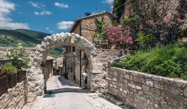 Una strada tranquilla in un borgo storico, con un arco in pietra e fiori colorati lungo il percorso.