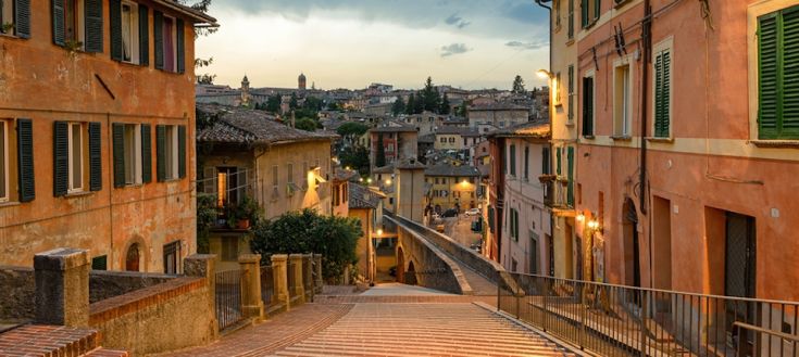 Una strada di città tranquilla al crepuscolo, con palazzi storici e luci che si accendono.