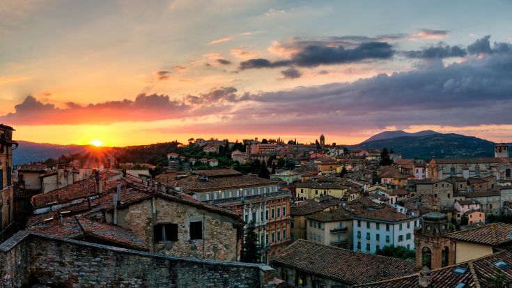 Panorama del tramonto su una città storica con palazzi dai tetti di pietra, circondata da dolci colline.