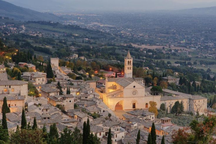 Panorama di Assisi al crepuscolo, con la basilica illuminata e le dolci colline circostanti.