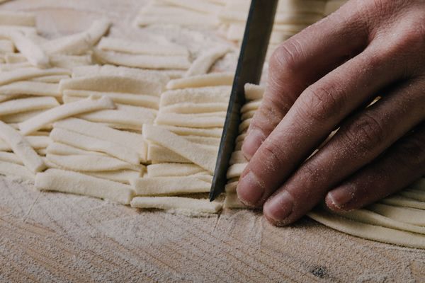 Un primo piano di una mano che taglia della pasta fresca su un piano di lavoro infarinato.