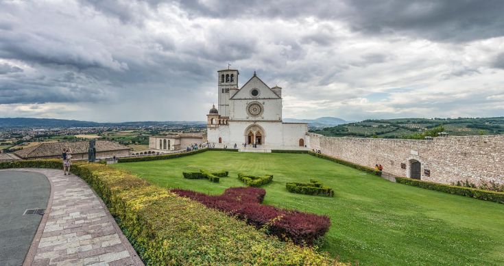 Panorama della Basilica di San Francesco, circondata da colline verdi e nuvole scure.