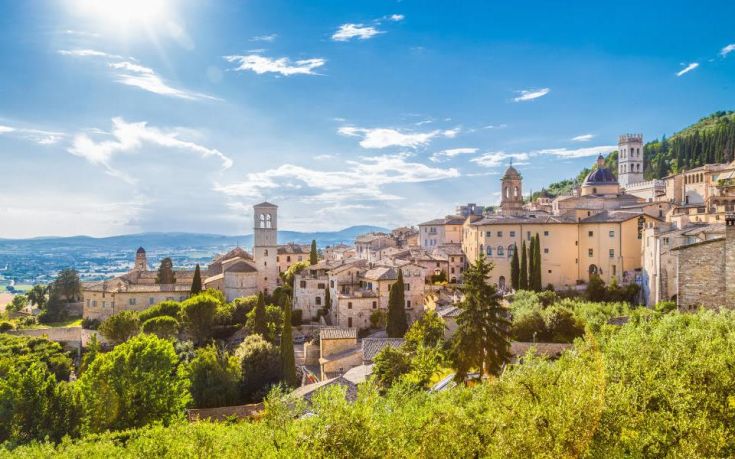 Scena tranquilla di Assisi circondata da colline, con edifici storici e un cielo azzurro.