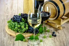 Due bicchieri di vino accanto a grappoli di uva verde e nera, sullo sfondo una botte in legno.