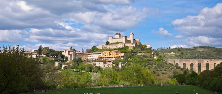 Rocca di Spoleto situata tra colline verdi, affiancato da vegetazione lussureggiante e velato da nuvole leggere.