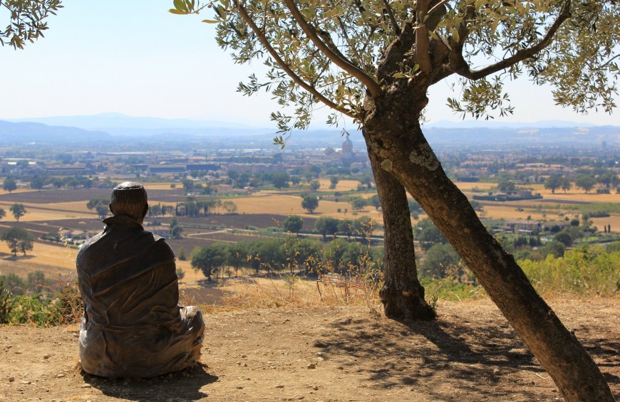 Una statua di ferro battuto in meditazione si affaccia sulla valle Umbra, con un albero d'ulivo accanto.