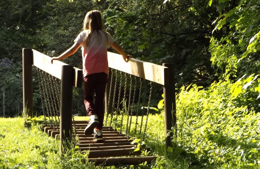 Una bambina attraversa un ponte in legno circondato da una natura verde e rigogliosa.