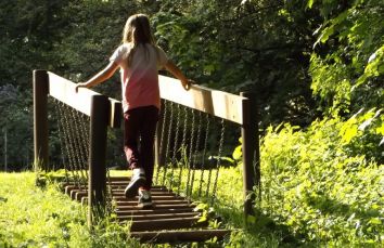 Una bambina attraversa un ponte in legno circondato da una natura verde e rigogliosa.