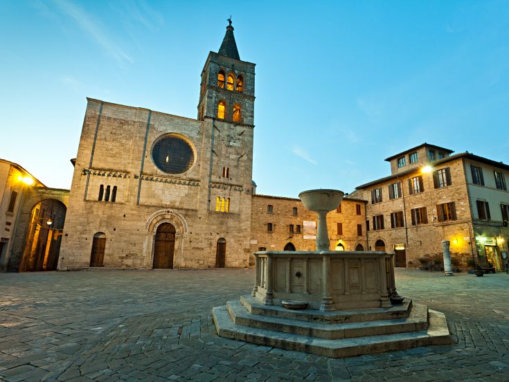 La piazza di Bevagna con la fontana e chiesa al tramonto, un'immagine sobria di architettura storica.
