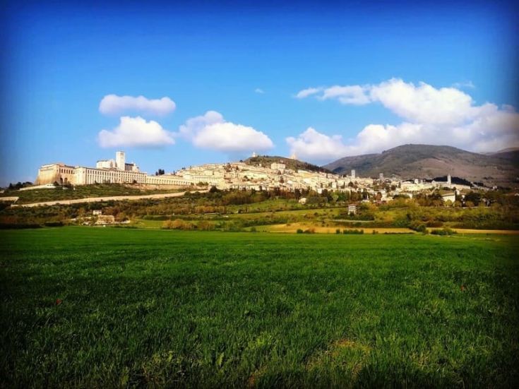 Scena di Assisi, circondata da campi verdi e un cielo sereno, che mette in risalto l'architettura del palazzo.