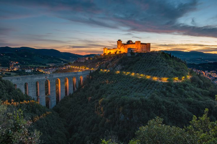 Spoleto illuminato al tramonto con il ponte delle due torri, circondato da colline verdi e un cielo blu. Una scena suggestiva.
