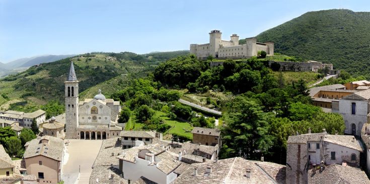 Panorama di un borgo storico con una chiesa e un antico palazzo, circondato da colline verdi.