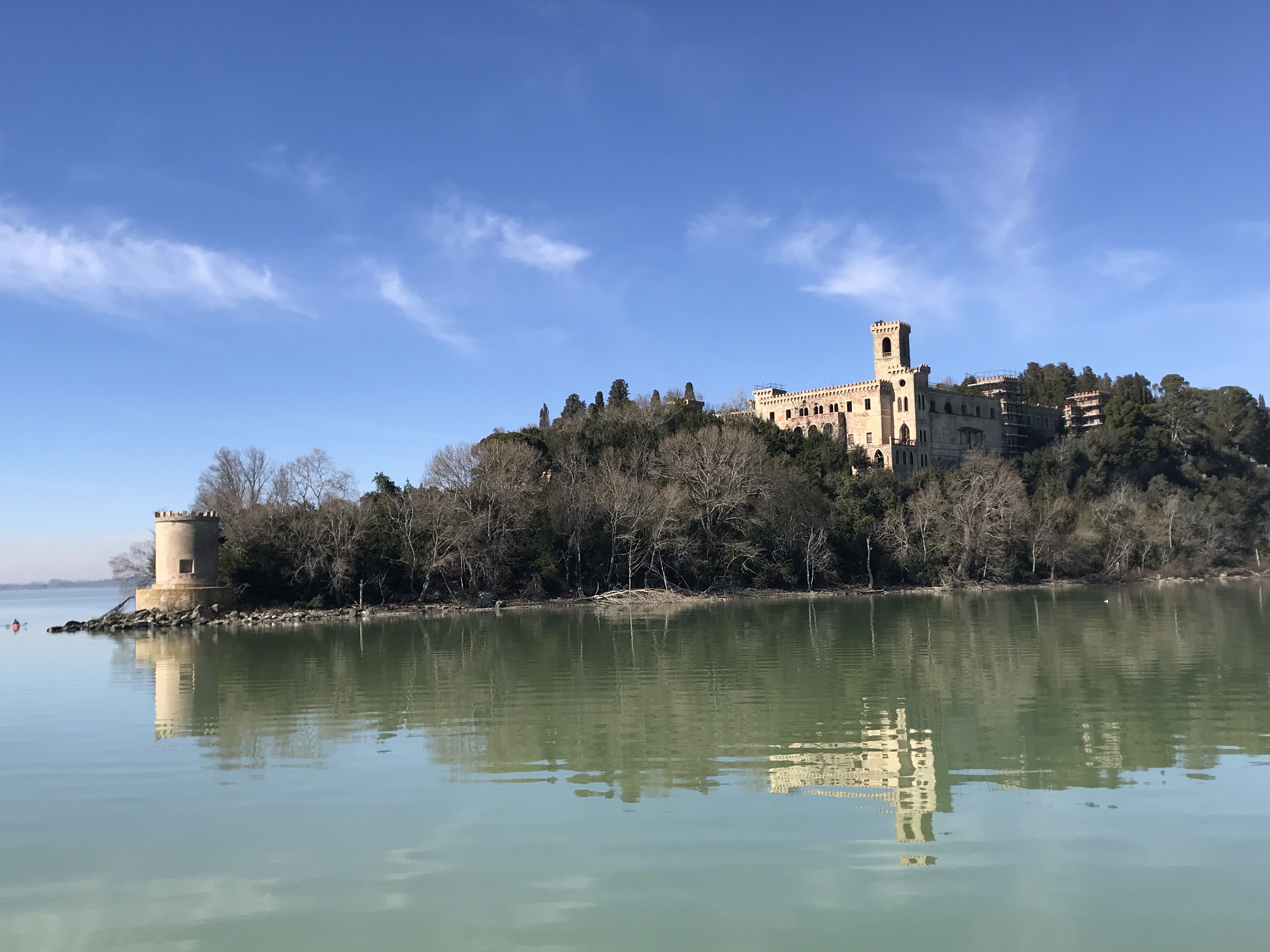 Un castello si riflette nel lago Trasimeno, circondato da alberi e vegetazione.