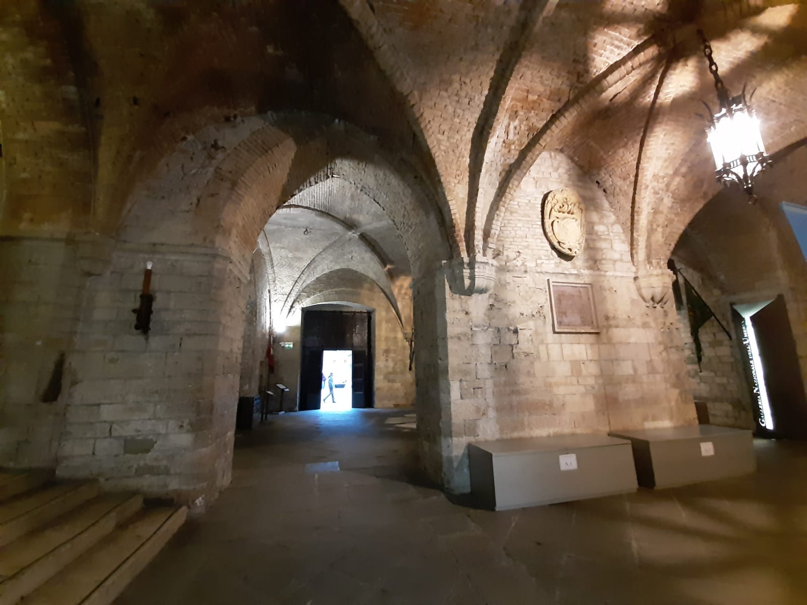 Galleria Nazionale dell'Umbria, ingresso con archi e luce naturale che entra da una porta, suggerendo un'atmosfera di storia e curiosità.