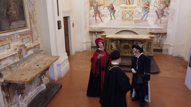 Un gruppo di persone vestite con costumi storici si trova in un palazzo decorato con affreschi, mentre intrattiene una conversazione.
