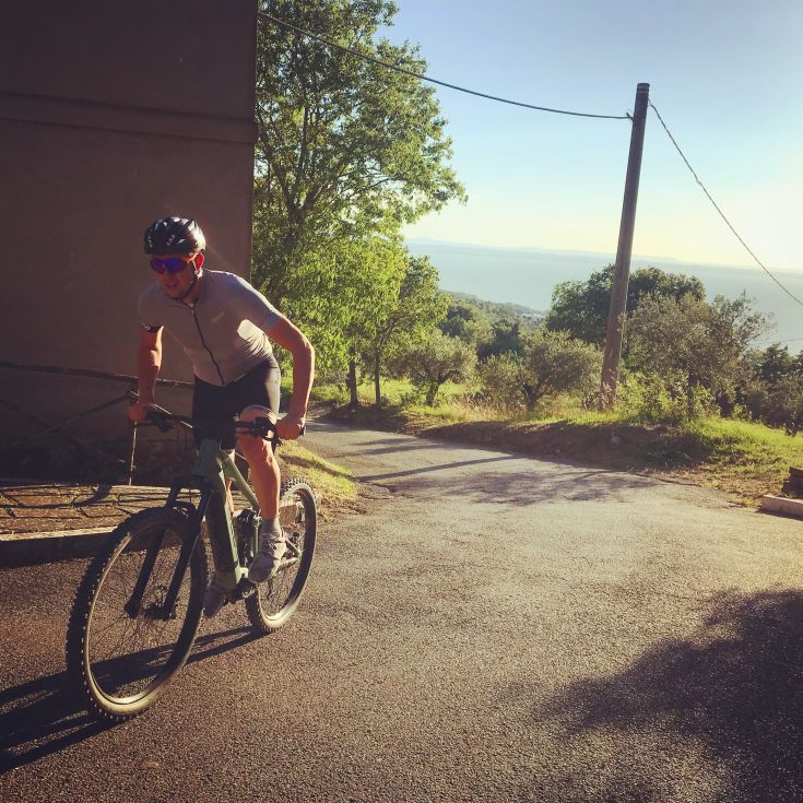 Un ciclista in movimento su una strada tranquilla, circondata da vegetazione. Il sole illumina il percorso mentre pedala sulla sua mountain bike.
