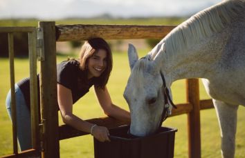 Una donna sorridente interagisce con un cavallo bianco mentre si nutre in un ambiente tranquillo.