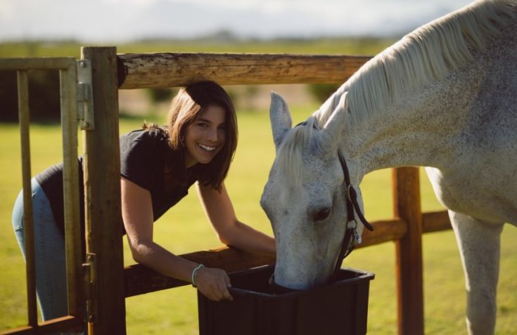 Una donna sorridente interagisce con un cavallo bianco mentre si nutre in un ambiente tranquillo.