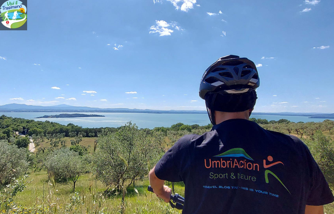 Un ciclista osserva il Lago Trasimeno da un punto panoramico, circondato da vegetazione e cielo sereno.