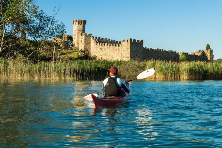 Un canoista si muove lentamente su un lago tranquillo, davanti a un palazzo immerso nella natura circostante.