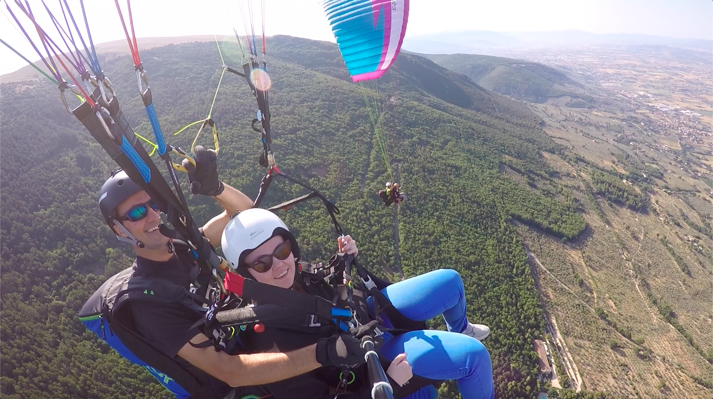 Due persone sorridenti mentre praticano parapendio sopra un paesaggio naturale.