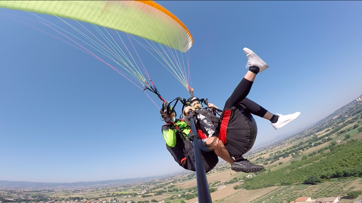 Due persone si librano in aria mentre praticano il parapendio, con una vasta area verde sottostante.