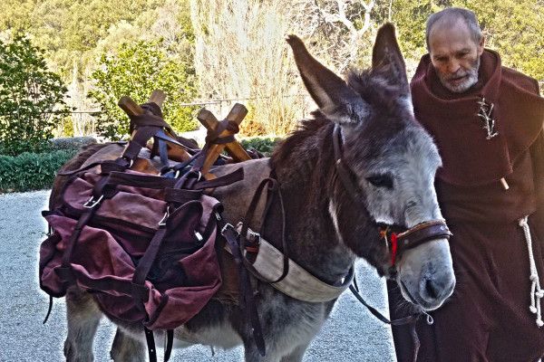Un uomo vestito da frate con un asino carico di sacchi in un contesto naturale. L'immagine rappresenta un momento legato alla tradizione agricola.